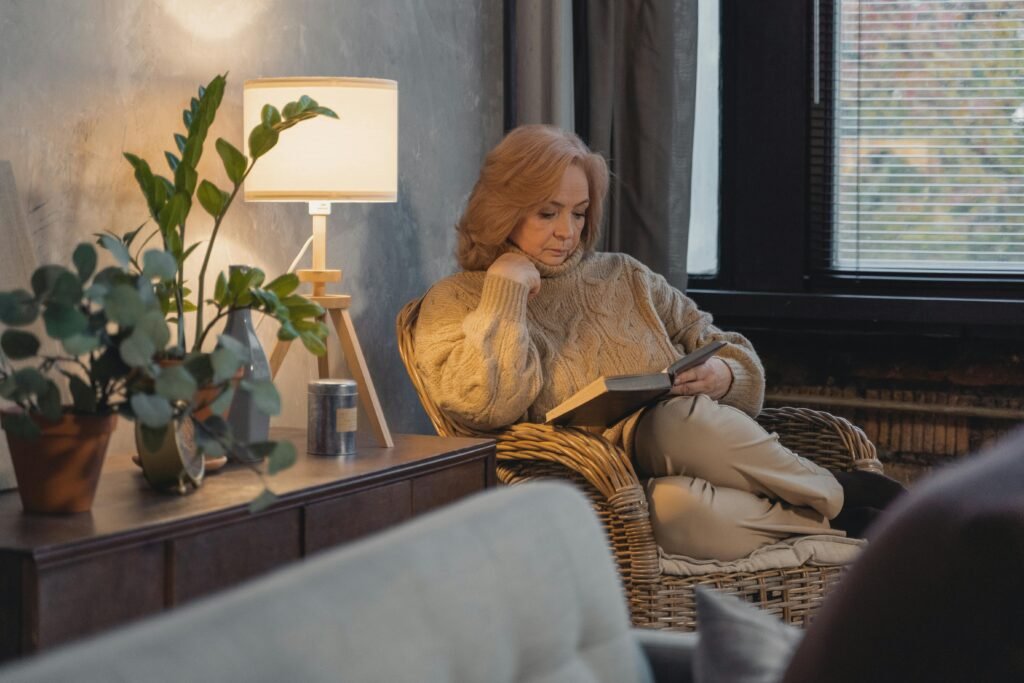 Senior woman in a sweater reading in a cozy room with plants and a lamp.