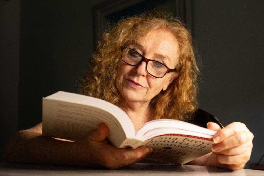 Senior woman with glasses reading a book in a cozy indoor setting, focused and relaxed.